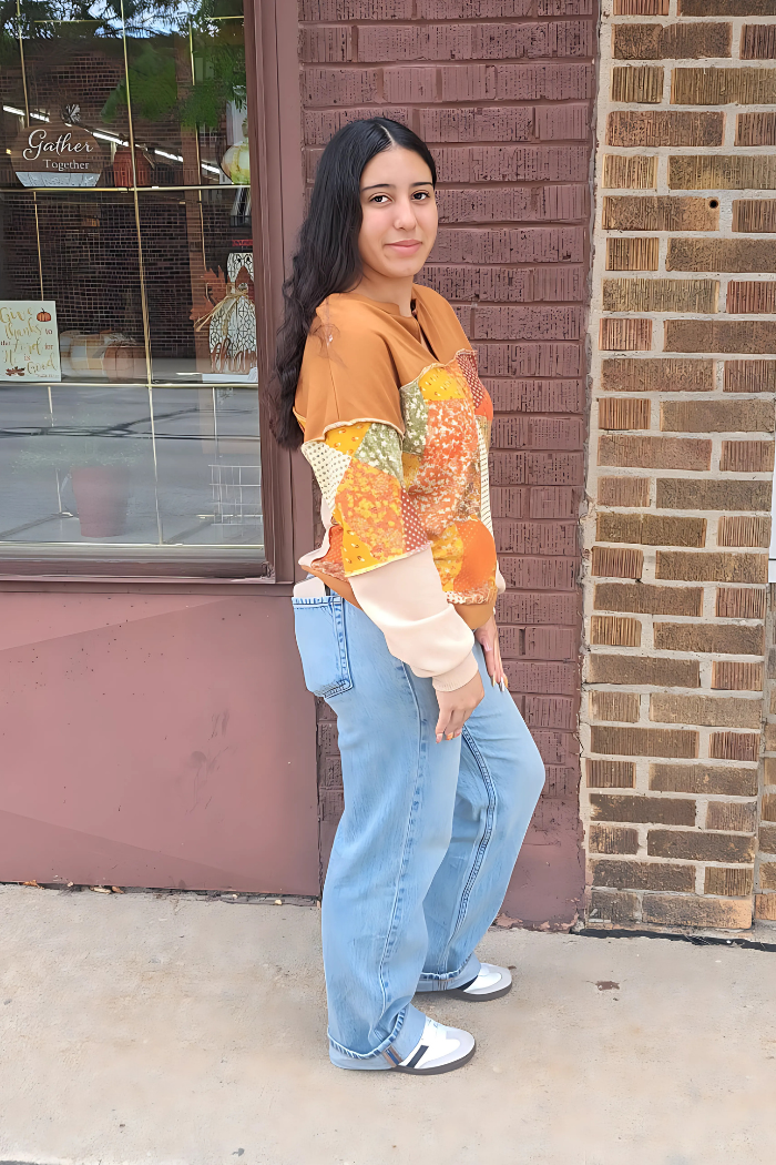 A young woman with dark hair stands on a sidewalk in front of a brick building with a large window. She is wearing a long-sleeved, earth-toned patchwork top and light blue jeans. Her hands are clasped in front of her, and she is looking towards the viewer with a slight smile. The window behind her displays decor and text that reads "Gather Together.