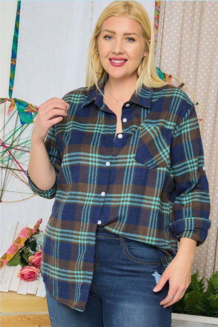 A smiling blonde woman wearing a blue, green, and brown plaid flannel shirt and blue jeans, standing indoors with a patterned curtain and colorful decorations in the background.