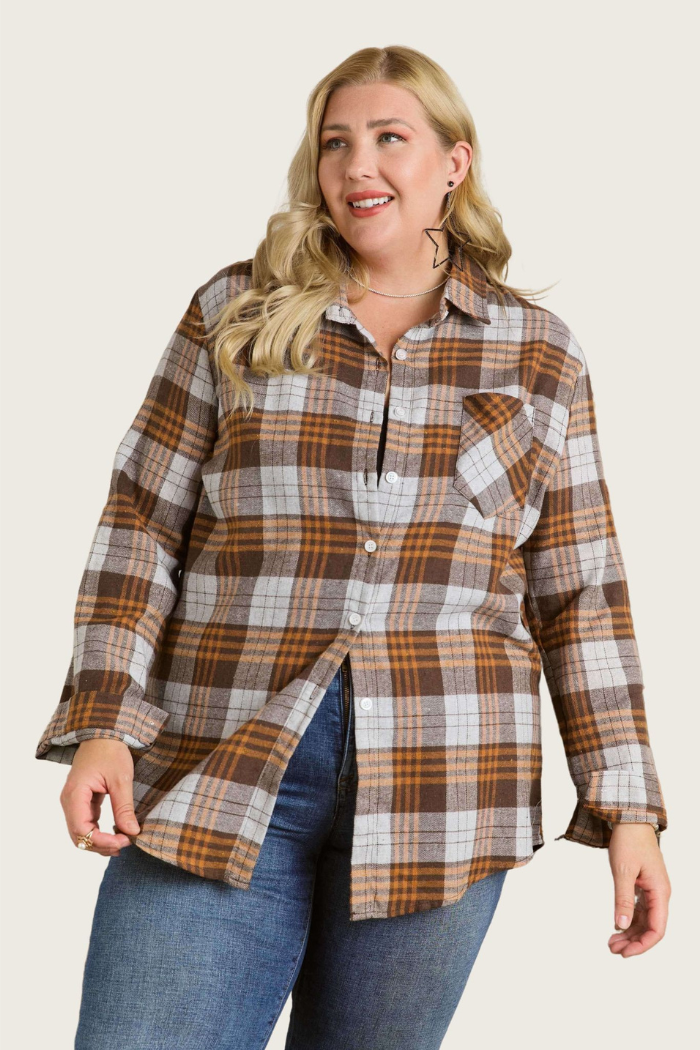 A plus-size woman with blonde hair smiles while posing in a brown/gray plaid flannel shirt. The shirt features a button-down front, long sleeves, and a single chest pocket on the left side. She is also wearing blue jeans. The background is a plain, light beige color. 