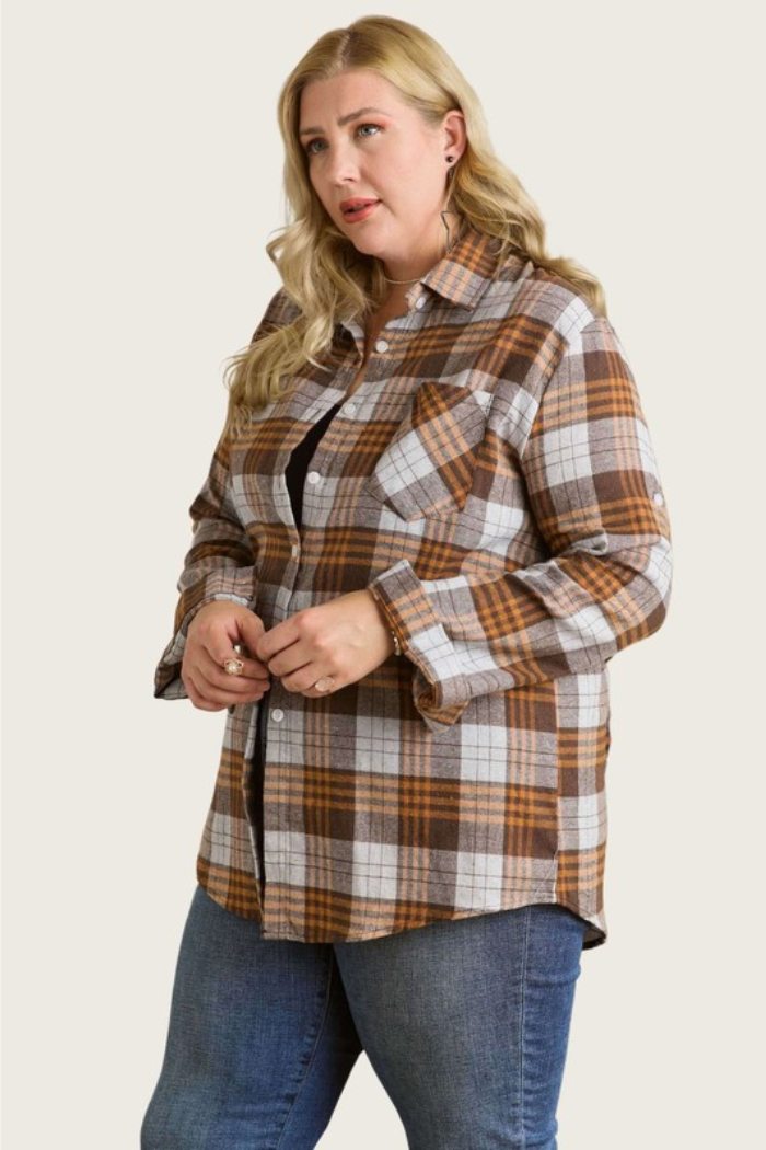 A blonde woman in a brown/grey plaid flannel shirt, unbuttoned to reveal a black top underneath, stands against a light beige background. She is looking slightly to her right, with her hands clasped in front of her. The shirt features a collar and a chest pocket.