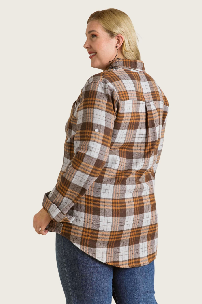A woman with blonde hair, wearing a brown/grey plaid flannel shirt, stands with her back mostly to the viewer, looking over her right shoulder and smiling. Her left arm is slightly bent, and her hand is near her hip. The shirt has long sleeves rolled up and secured with a tab and button. She is wearing dark blue jeans. The background is a plain, light, solid color.