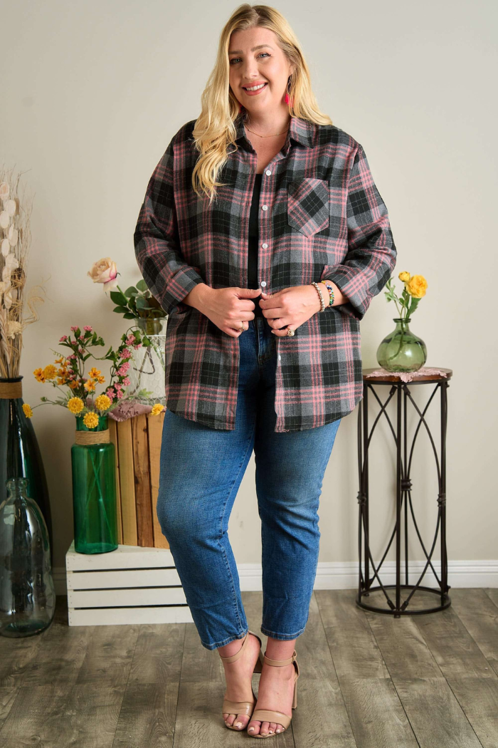 A smiling plus-size woman with long blonde hair stands in a room, wearing a grey/black and pink plaid flannel shirt over a black top, blue jeans, and light brown open-toed heels. She is holding the bottom of her shirt closed with both hands. The background features various decorative elements including large glass bottles, flowers in vases, and a small table with a metal base. 