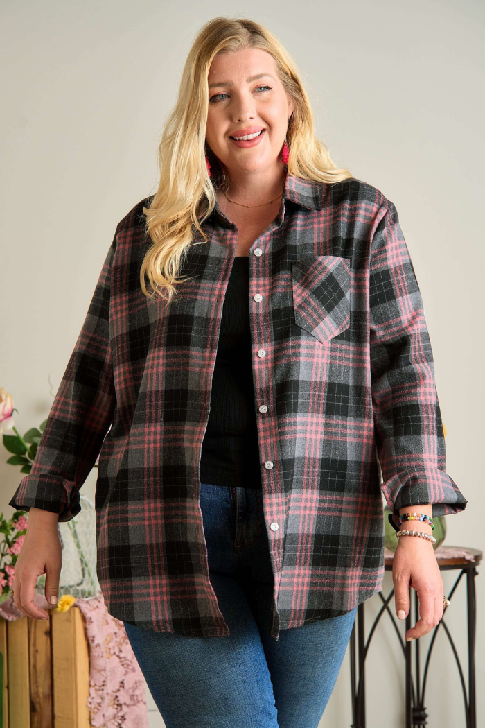 A smiling blonde woman with long hair wears an open, oversized, long-sleeved plaid flannel shirt in shades of pink, grey, and black over a black top and blue jeans. She is standing and looking directly at the camera. The setting appears to be indoors with a light-colored background and some subtle floral decorations visible on the left.