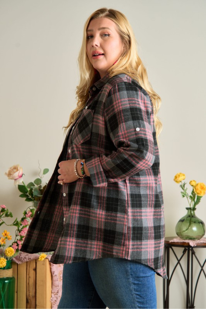 A plus-size woman with long blonde hair wears a gray/black and pink plaid flannel shirt over blue jeans. She is standing and looking towards the viewer, with her body slightly turned to the side. She has several bracelets on her left wrist. The background is a light-colored wall with some decorative elements, including what appears to be a plant with pink flowers on the left.
