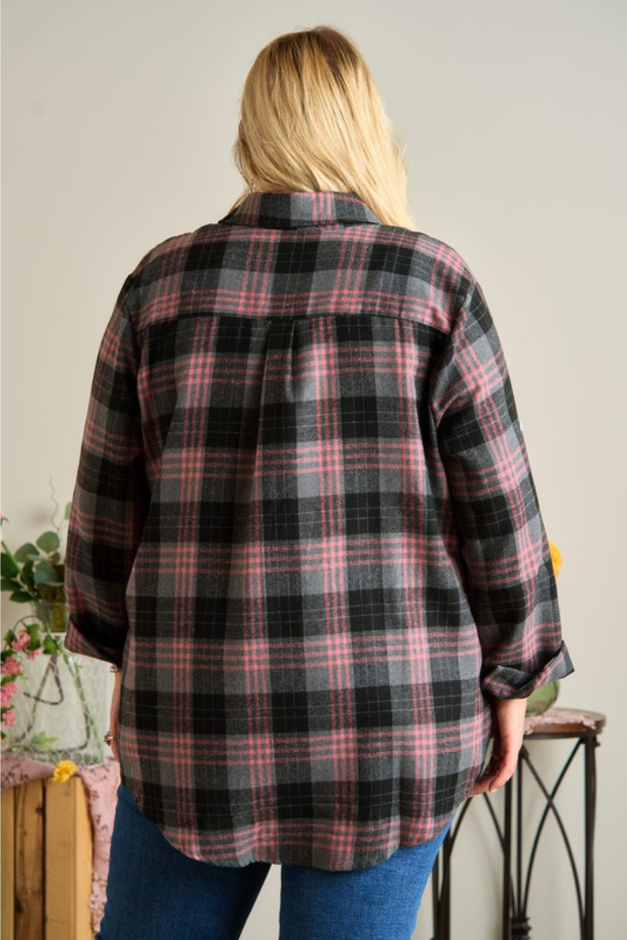 A woman from the back, wearing a loose-fitting, long-sleeved plaid flannel shirt in shades of black, grey, and pink. The shirt has a rounded hem and the sleeves are rolled up. She is also wearing blue jeans.