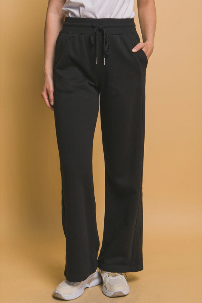 A person stands, facing forward, wearing black wide-leg sweatpants with a visible drawstring at the waist and pockets. The sweatpants are paired with a light-colored top and light-colored athletic shoes.