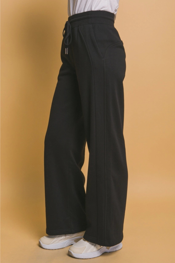 A person stands, sideways wearing black wide-leg sweatpants with a visible drawstring at the waist and pockets. The sweatpants are paired with a light-colored top and light-colored athletic shoes.