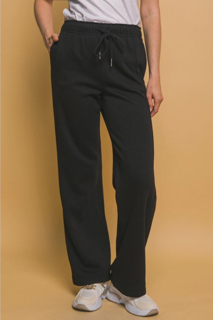 A person standing against a plain light brown background, wearing black wide-leg sweatpants with a visible drawstring at the waist, a plain white t-shirt, and white sneakers with tan accents. One hand is in the pocket of the sweatpants.