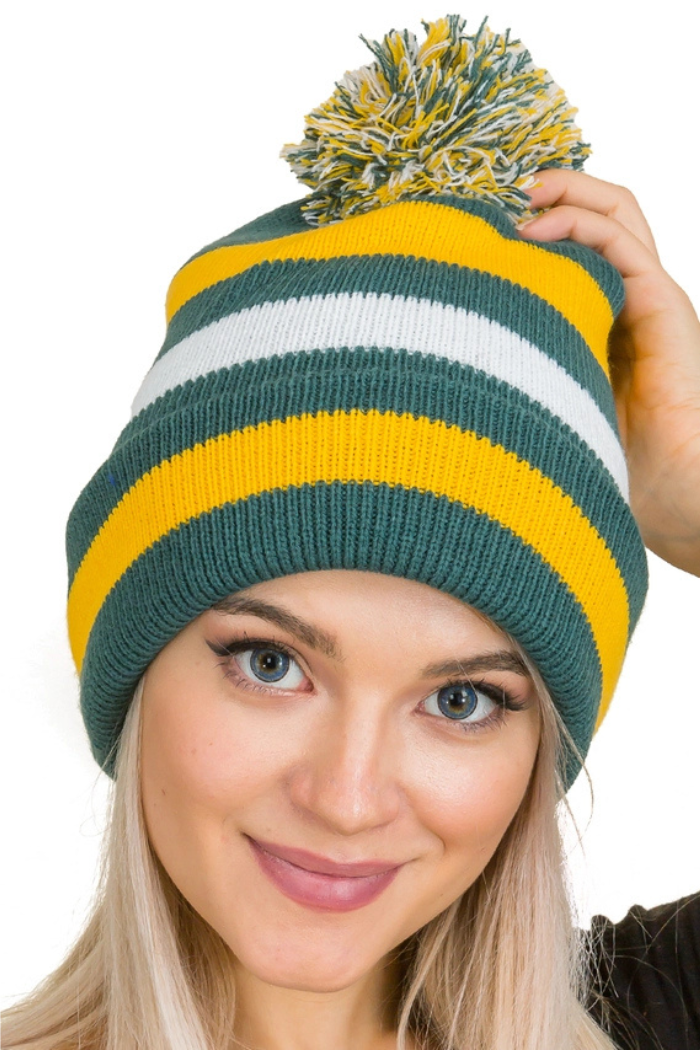 A close-up shot features a person wearing a cuffed knit beanie with a pom-pom on top. The beanie is primarily green, white and yellow with a prominent white stripe across the cuff. The person's face is visible, with light-colored hair and blue eyes, and they are looking directly at the viewer with a slight smile. The background is solid white.