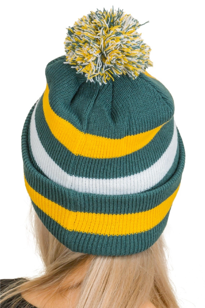 A knitted winter hat (beanie) viewed from the back, worn by a person with blonde hair. The hat is primarily dark green with horizontal stripes in yellow and white. It features a large, fluffy pom-pom on top, composed of green, yellow, and white threads. 