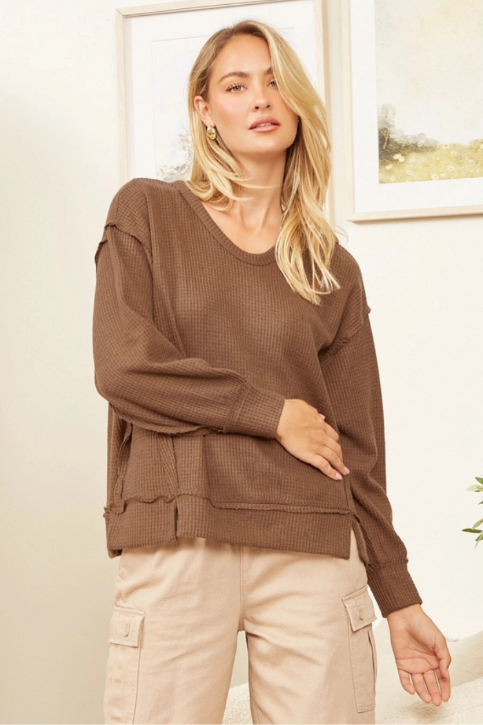A woman with blonde hair wearing a brown waffle-knit long-sleeve top and light-colored cargo pants stands in a room with framed artwork on the wall. The top has a round neck and distressed details on the seams and hem. The woman is looking directly at the camera with a slight smile.