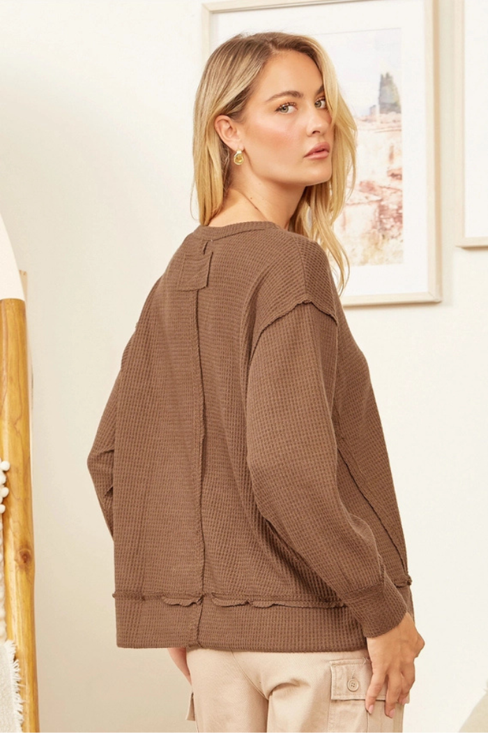 A woman with blonde hair wearing a brown waffle-knit long-sleeve top with exposed seams, looking over her right shoulder, with a blurred background featuring a mirror and framed art.