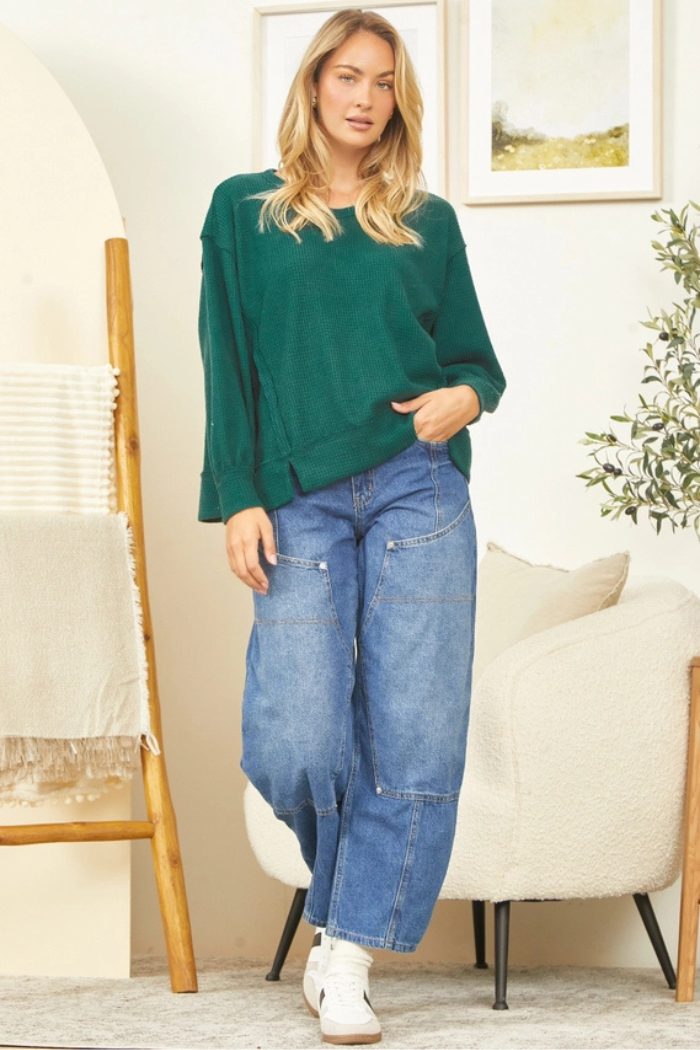 A woman with long blonde hair stands casually in an indoor setting, wearing a dark green, relaxed-fit top and light blue, wide-leg or "barrel" style jeans with visible seam details. She is also wearing white sneakers. Her left hand is in her pocket, and she is looking directly at the viewer with a slight smile. In the background, there's a light-colored ladder-style shelf with blankets on the left, and a cream-colored armchair and a potted plant with olive-like leaves on the right.