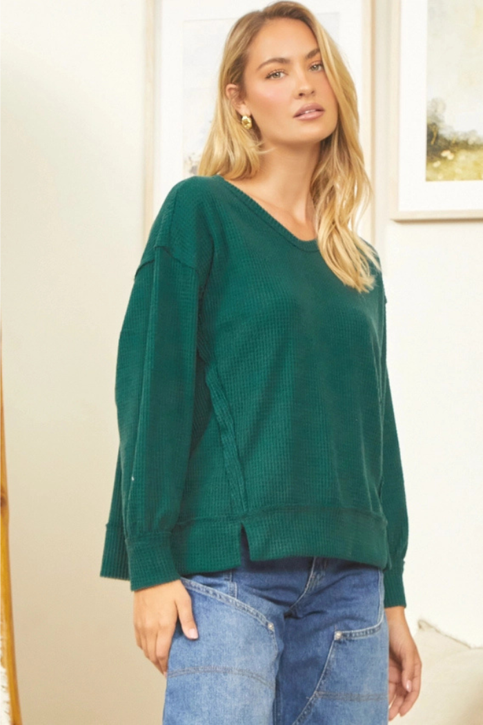 A woman with blonde hair wearing a dark green, oversized, round neck waffle knit long-sleeved top and blue jeans, standing indoors.
