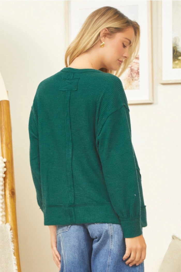 A woman with blonde hair, facing away from the viewer and slightly to the right, wears a dark green, long-sleeved, oversized waffle-knit top and blue jeans. She is standing indoors, with a framed picture visible on the wall behind her.