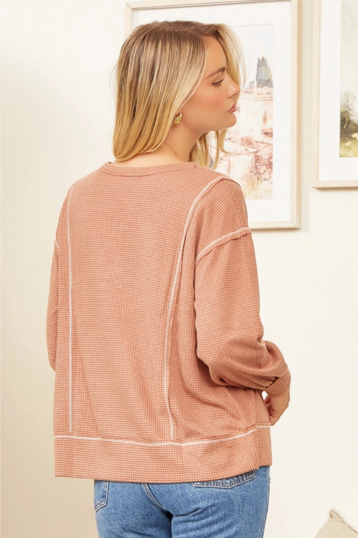 A woman seen from the back, with blonde hair, wears a long-sleeved, rust-colored or peach-toned thermal waffle-knit top with prominent white contrast stitching details along the seams of the sleeves and sides. She is also wearing blue jeans. The background appears to be an indoor setting with framed artwork on the wall.