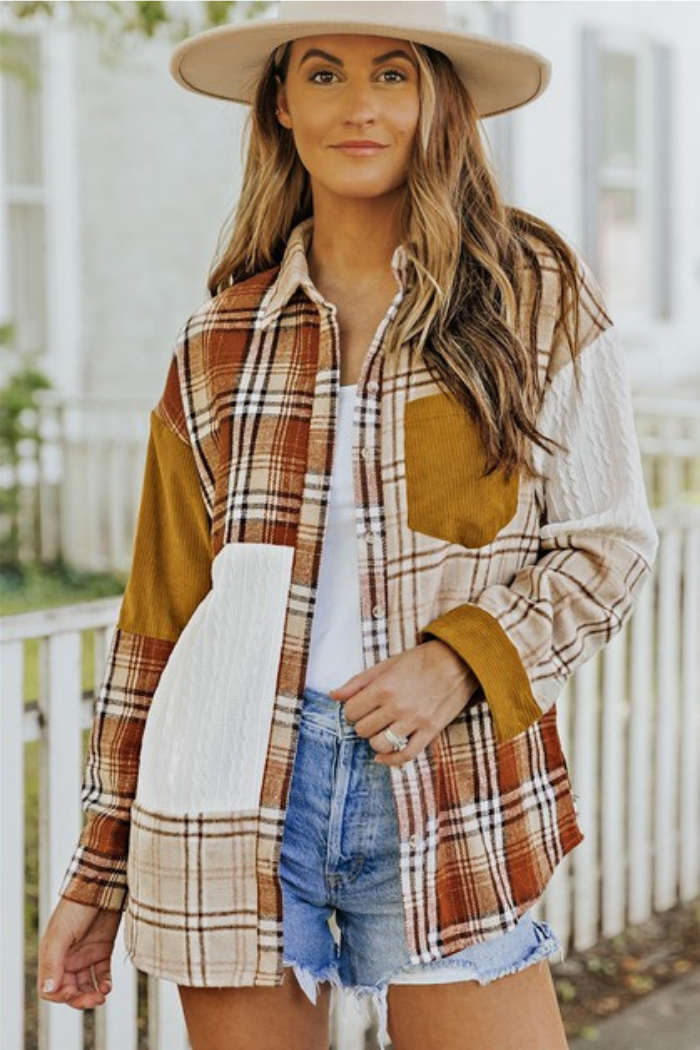 A woman wearing a beige fedora hat, patchwork plaid shirt jacket with a mix of brown, beige, and white plaid patterns, along with solid brown corduroy and textured white fabric patches. The jacket has a button-down front and a pocket on the left chest area. She is also wearing a white top underneath and blue shorts. She is smiling and looking at the camera.
