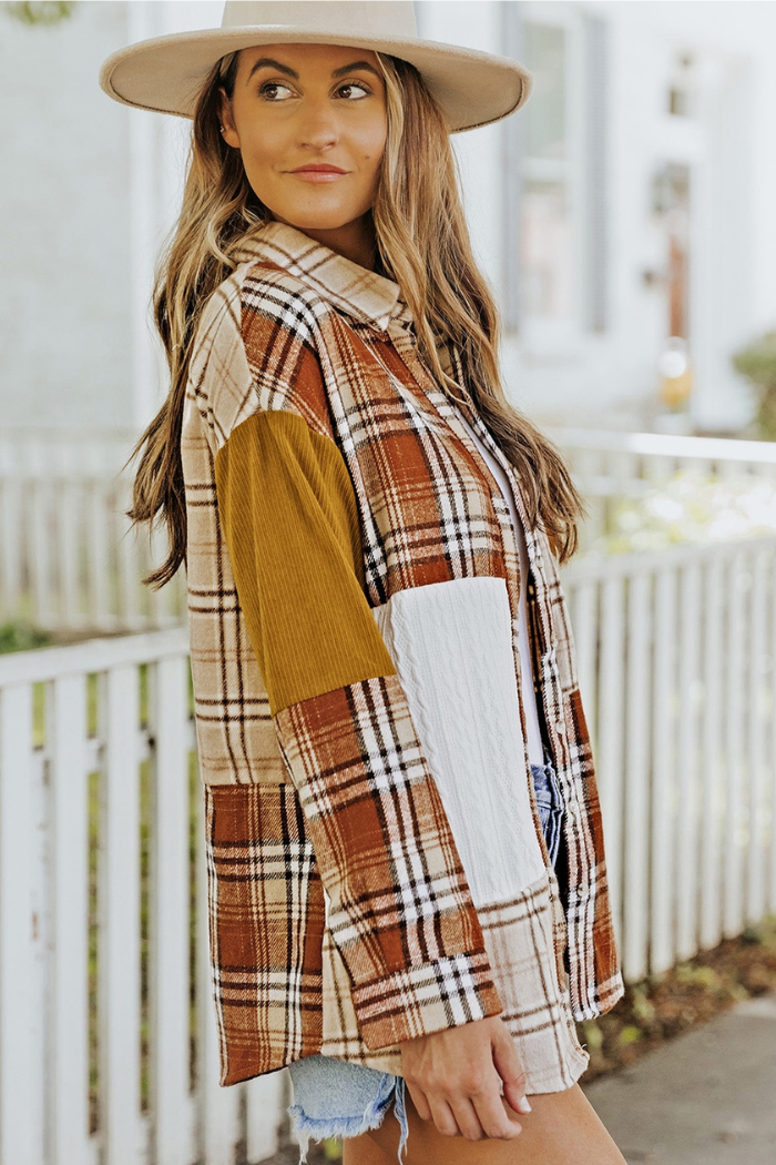 A woman wearing a beige hat and a plaid color-block patchwork shirt jacket, featuring various shades of brown, beige, and white plaid patterns, with a solid mustard yellow corduroy section on the left sleeve and a textured white panel on the right side of the torso. She is looking to her right and slightly smiling, with long, wavy brown hair. The background appears to be an outdoor setting with a light-colored fence or railing behind her.