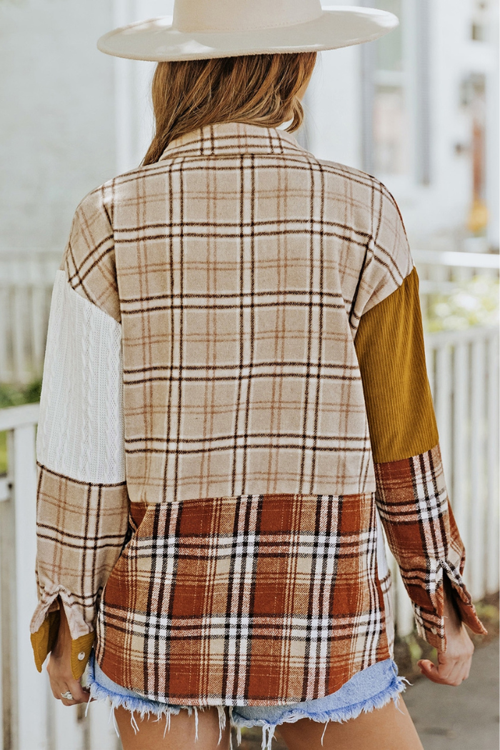 A woman wearing a beige and brown plaid color-block patchwork shirt jacket with a white cable knit sleeve on the left and a solid brown corduroy sleeve on the right, viewed from the back. She is also wearing a wide-brimmed hat and denim shorts. The background shows a white fence and greenery.