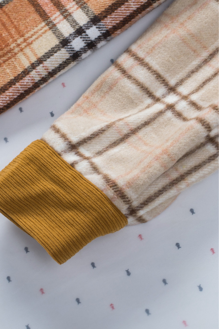 A close-up of a beige and brown plaid shirt jacket with a ribbed, mustard-colored cuff on the sleeve. The plaid pattern features varying shades of beige, brown, and a hint of orange.