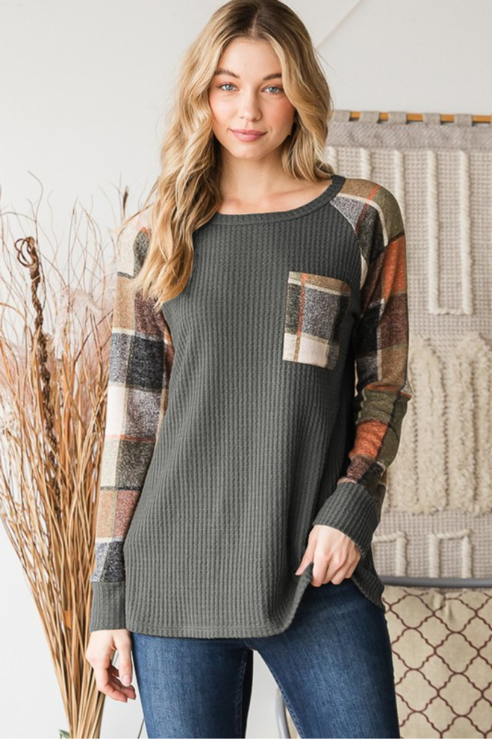 A woman with long, blonde hair models a long-sleeved, waffle-knit top in a charcoal grey color. The top features raglan sleeves and a single chest pocket, both made from a plaid fabric in shades of brown, tan, and orange. She is wearing blue jeans and standing in front of a textured background with natural elements on the left.