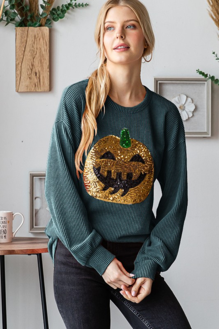 A woman wearing a green, ribbed long-sleeved top with a large, gold sequined jack-o'-lantern design on the front. The jack-o'-lantern has black features and a green sequin stem. She is also wearing dark pants. 
