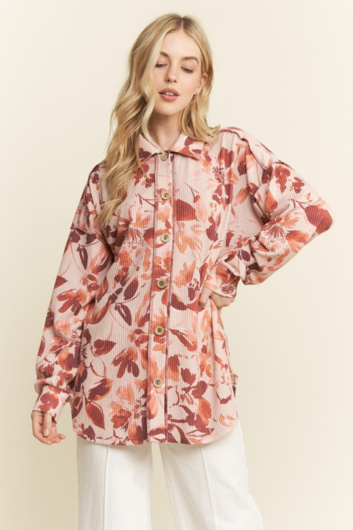 A blonde woman models a long-sleeved, collared button-down shirt with a floral pattern in shades of pink, orange, and taupe. The shirt appears to be made of a ribbed or corduroy-like fabric and is worn over white pants. The woman is standing with her hands on her hips, looking forward.