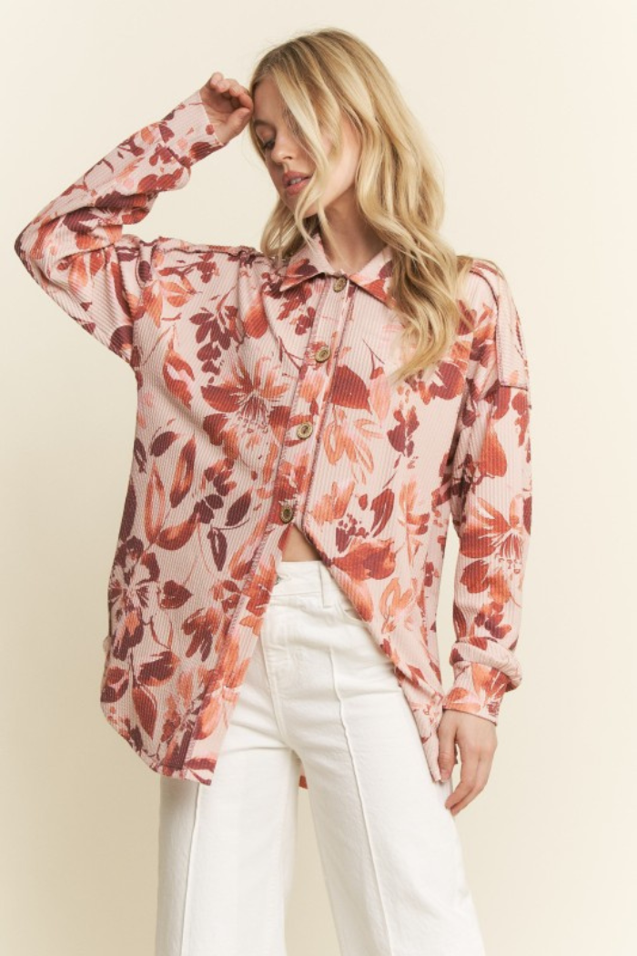 A blonde woman wearing a light pink, long-sleeved, collared button-down shirt with a floral pattern in shades of pink, orange, and taupe, paired with white wide-leg pants. She is standing with one hand raised to her head, looking downwards.
