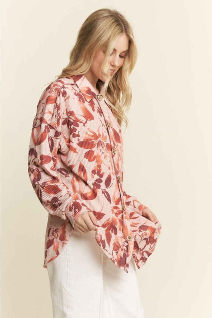 A blonde woman with long hair wearing an oversized button-down shirt with a taupe and orange floral print, paired with white pants. She is standing and looking down, holding the bottom hem of the shirt with both hands. 