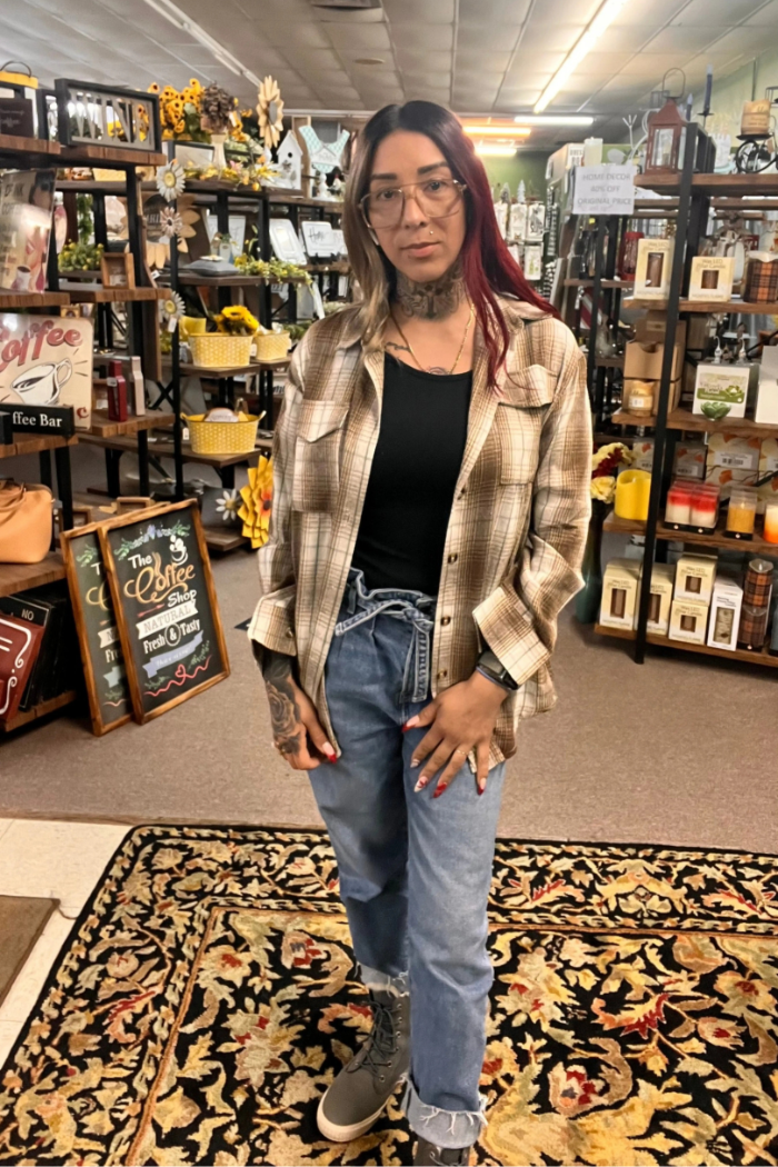 A person stands centered in a store, wearing a brown and beige plaid flannel shirt over a black top, light wash jeans with a high ruffled waist, and gray sneakers. The background shows shelves filled with various items, including decorative elements like sunflowers and lanterns, and a chalkboard sign for "The Coffee Shop." 