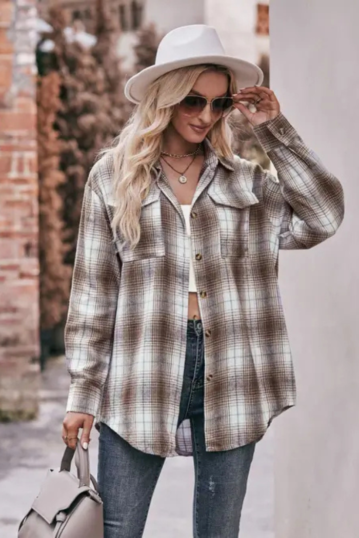 A woman stands outdoors, wearing a light-colored fedora-style hat and sunglasses. She has on a long-sleeved, oversized plaid shirt in shades of brown, white, and a hint of blue, layered over a white top. She is wearing blue jeans and carrying a light grey shoulder bag or purse on her left arm. The background suggests an urban or outdoor setting with a building and wall behind her.