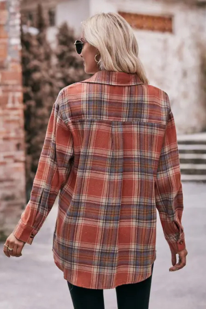 A person seen from behind, walking away, wearing an oversized long-sleeved plaid shirt in shades of orange, blue, and tan. They are wearing sunglasses and facing a brick building.