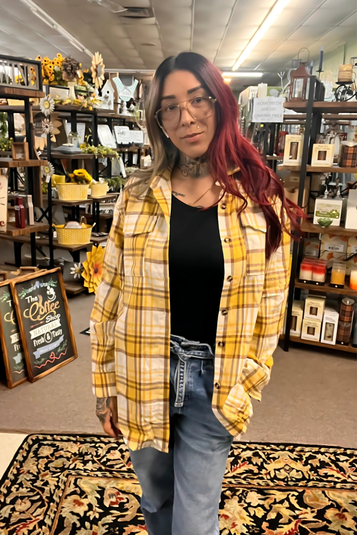 A woman wearing a yellow and white plaid shacket, a popular style that combines elements of a shirt and a jacket. She is wearing it unbuttoned over a plain black top, paired with blue jeans.