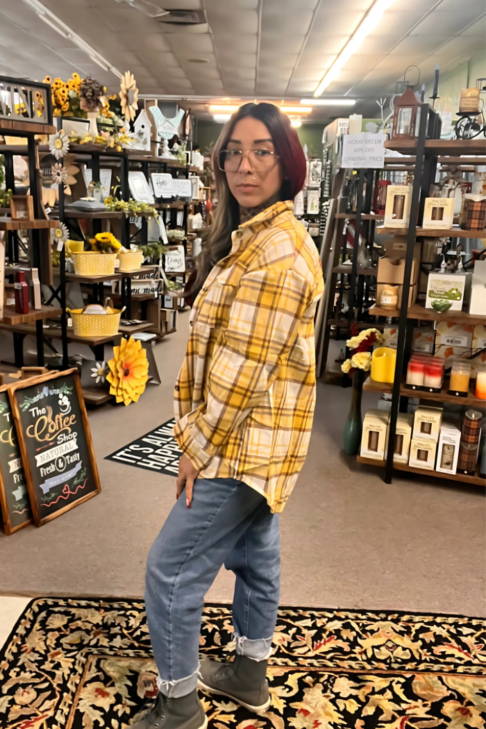 A woman with shoulder-length reddish-brown hair and glasses stands in a store, looking slightly to her left. She is wearing a yellow and white plaid button-up shirt, blue jeans, and dark shoes. The background shows shelves and displays of various merchandise, including home decor items and clothing.