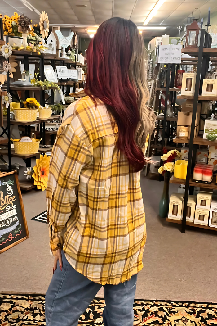 A person with long, two-toned hair (dark red on one side, light brown/blonde on the other) stands with their back to the viewer, wearing a loose-fitting yellow and white plaid button-down shirt and blue jeans, in what appears to be a retail store or boutique setting with various home decor items visible in the background.