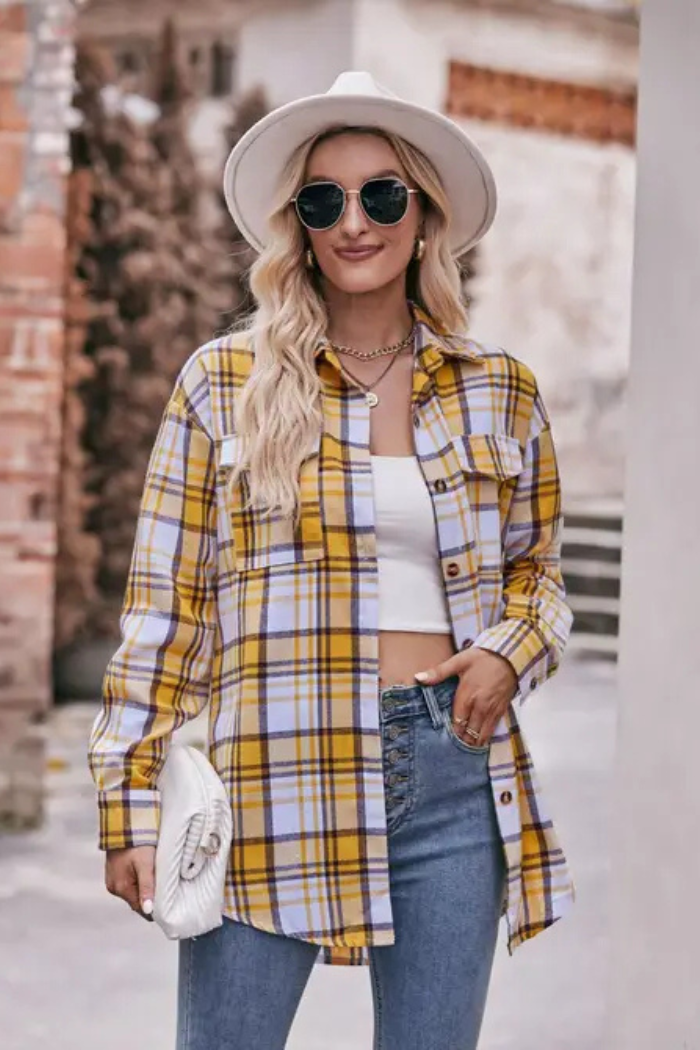 A woman wearing a yellow and white plaid long-sleeve button-down shirt over a white crop top, paired with blue jeans. She is also wearing a white wide-brim hat, sunglasses, and a gold-toned necklace, and is holding a white quilted handbag. 
