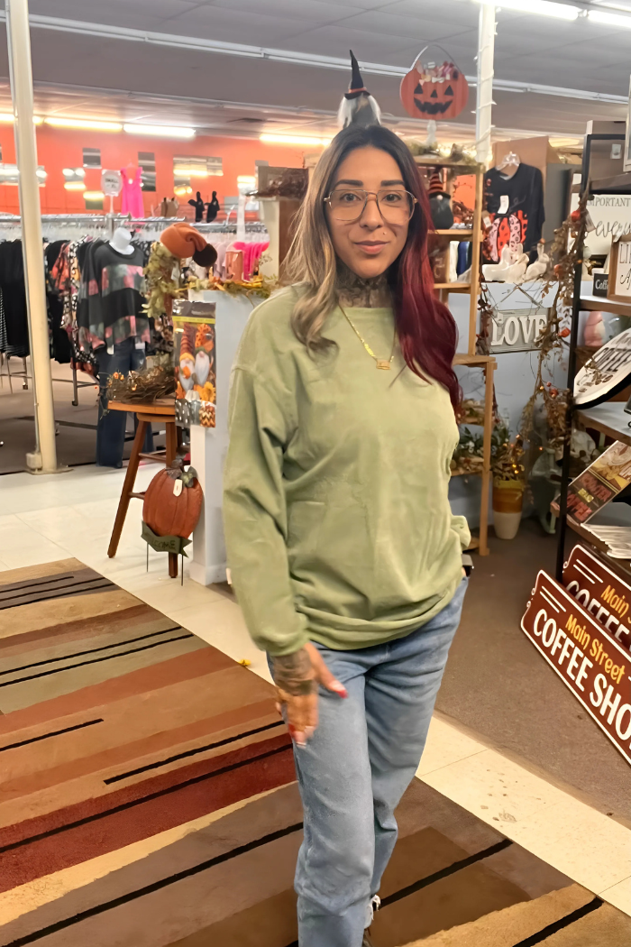 wearing an oversized, light olive-green long-sleeved sweatshirt and light blue jeans. In the background, there are clothing racks on the left and various fall or Halloween-themed decorations and signage on the right, including pumpkins and a sign that reads "Main Street COFFEE SHOP.
