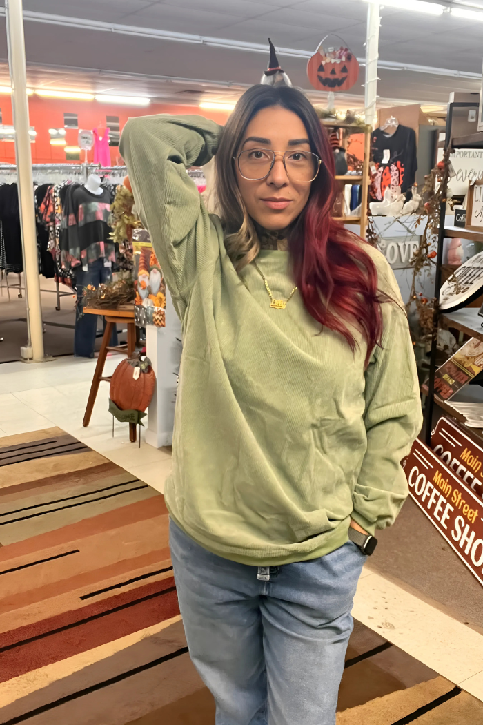 A woman with long, dark hair with red highlights is wearing an olive-green sweatshirt and light wash jeans. She is looking directly at the camera with a slight smile. Her right arm is raised with her hand resting on her head, and her left hand is in her pocket.