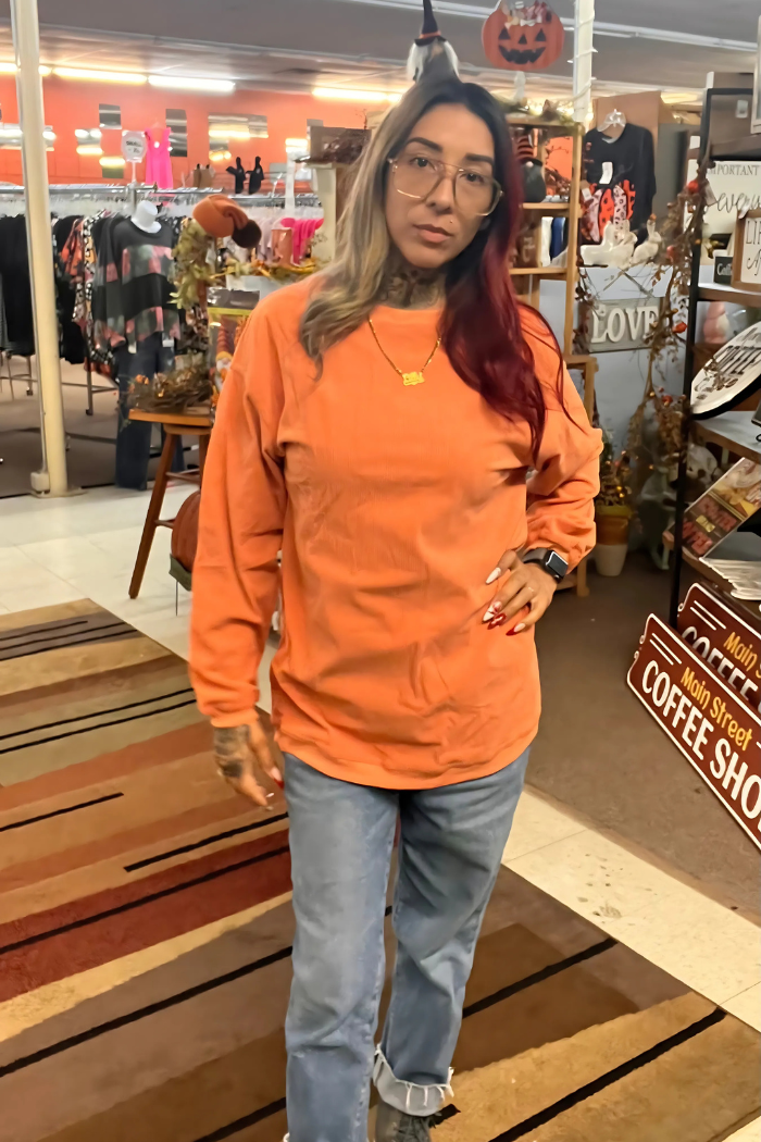 A woman with long hair, dyed half blonde and half reddish-brown, stands indoors wearing glasses, an orange ribbed long-sleeved shirt, and light-wash jeans. She is posing with her left hand on her hip.