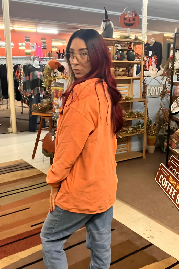 The image shows aside view person wearing a casual and cozy fall-inspired outfit, featuring a vibrant orange ribbed oversized sweatshirt paired with light-wash jeans.