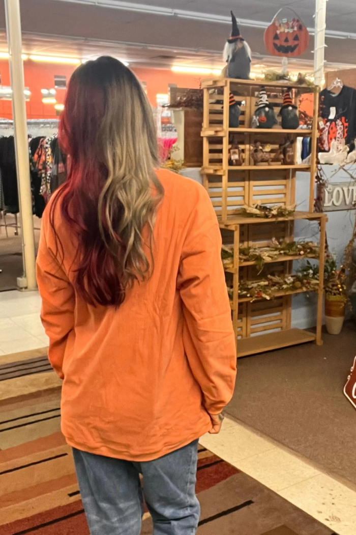 A person viewed from behind, wearing an orange ribbed long-sleeved sweatshirt and blue jeans, stands in what appears to be a store. The person has long hair that transitions from a darker color to a lighter, possibly dyed, shade.
