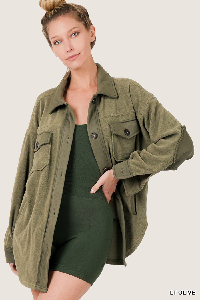 A woman with her hair in a bun models an oversized, light olive green fleece shacket over a dark olive green ribbed bodysuit. The shacket has a button-up front, two chest pockets, and elbow patches. She is standing with her left hand on her hip, looking towards the camera. 