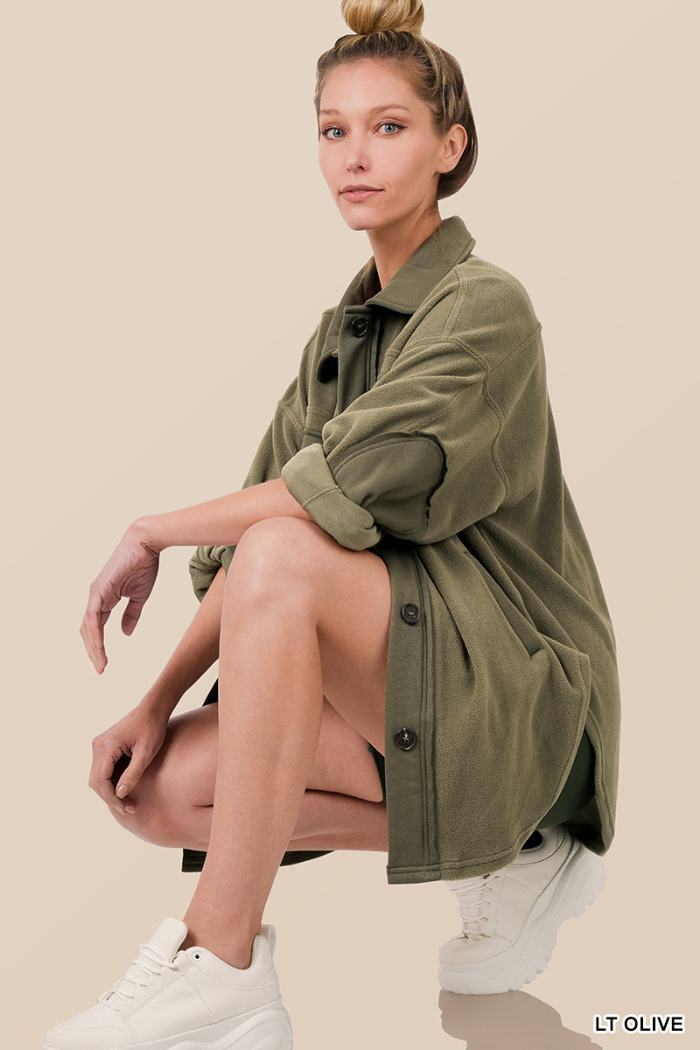 A woman with light hair in a high bun wears the Fleece Oversized Button Shacket in olive green, sitting crouched with one knee up, arms resting on her leg, styled with white sneakers against a beige background.