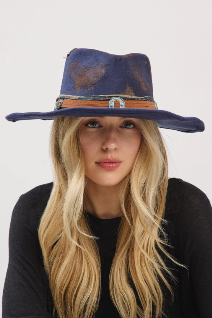 A woman with long blonde hair and a black top wears a distressed, dark blue felt fedora hat with a wide brim and a tan leather band featuring turquoise-colored accents.