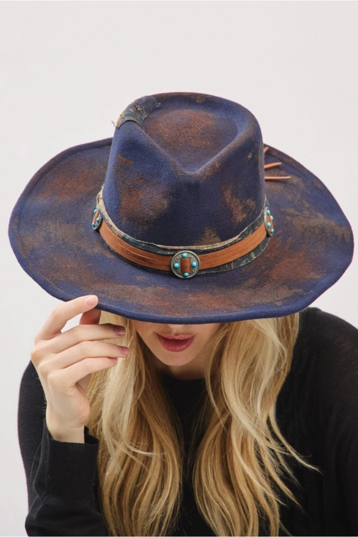 A young woman with long blonde hair holds the brim of a distressed, dark blue fedora-style hat with a wide brim, covering her eyes. The hat features a decorative band with turquoise-colored accents and a faux-suede texture. She is wearing a dark top, and the background is plain white.