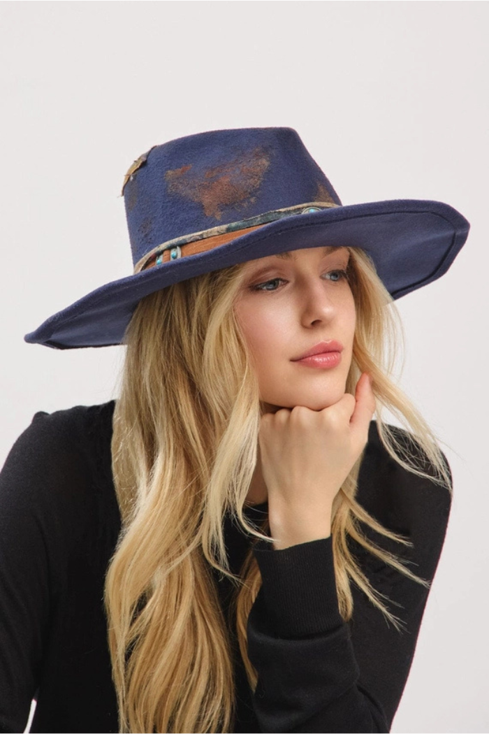 A blonde woman in a black top wears a dark blue, distressed-look cowboy or fedora-style hat with a decorative band and turquoise accents, looking thoughtfully to her right with her hand under her chin. 