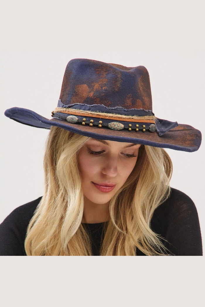 A woman with blonde hair wearing a distressed, dark blue wide-brimmed hat with brown and copper accents, a studded band, and decorative stones.