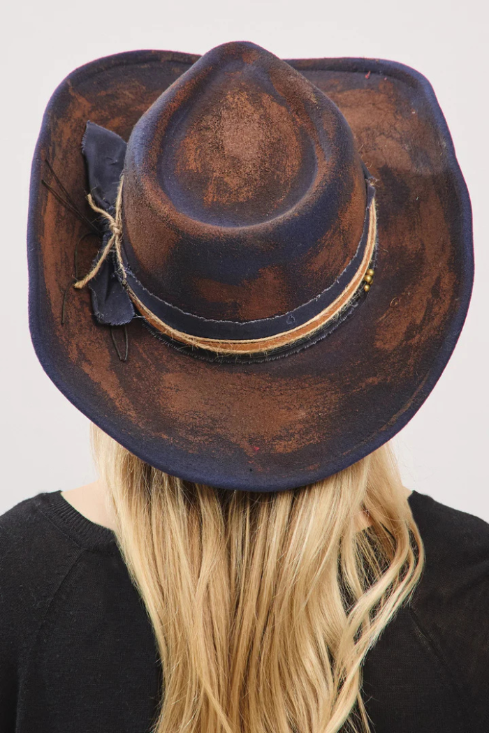 The image displays the back of a person with long, blonde hair, wearing a distressed dark blue and brown cowboy hat. The hat features a textured band with a bow and subtle embellishments.