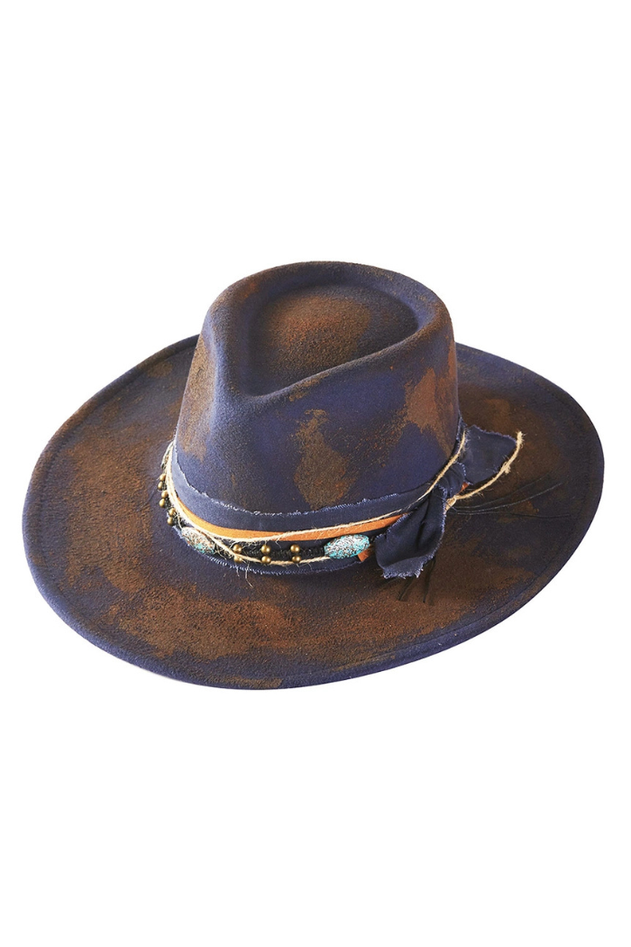 A distressed western-style fedora hat in dark blue with rustic brown aging accents, featuring a multi-layered, textured band adorned with beads and a fabric bow.