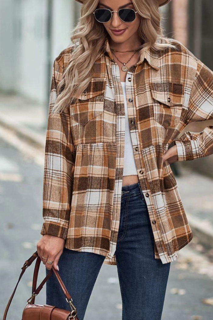 A stylish woman with long blonde hair wears a brown and tan plaid flannel shirt over a white top, paired with dark blue jeans. She stands outdoors, looking down and holding a brown shoulder bag in her left hand, with a street or path visible in the background.